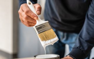 Painter's hand holding angled brush dripping white paint over open can indoors