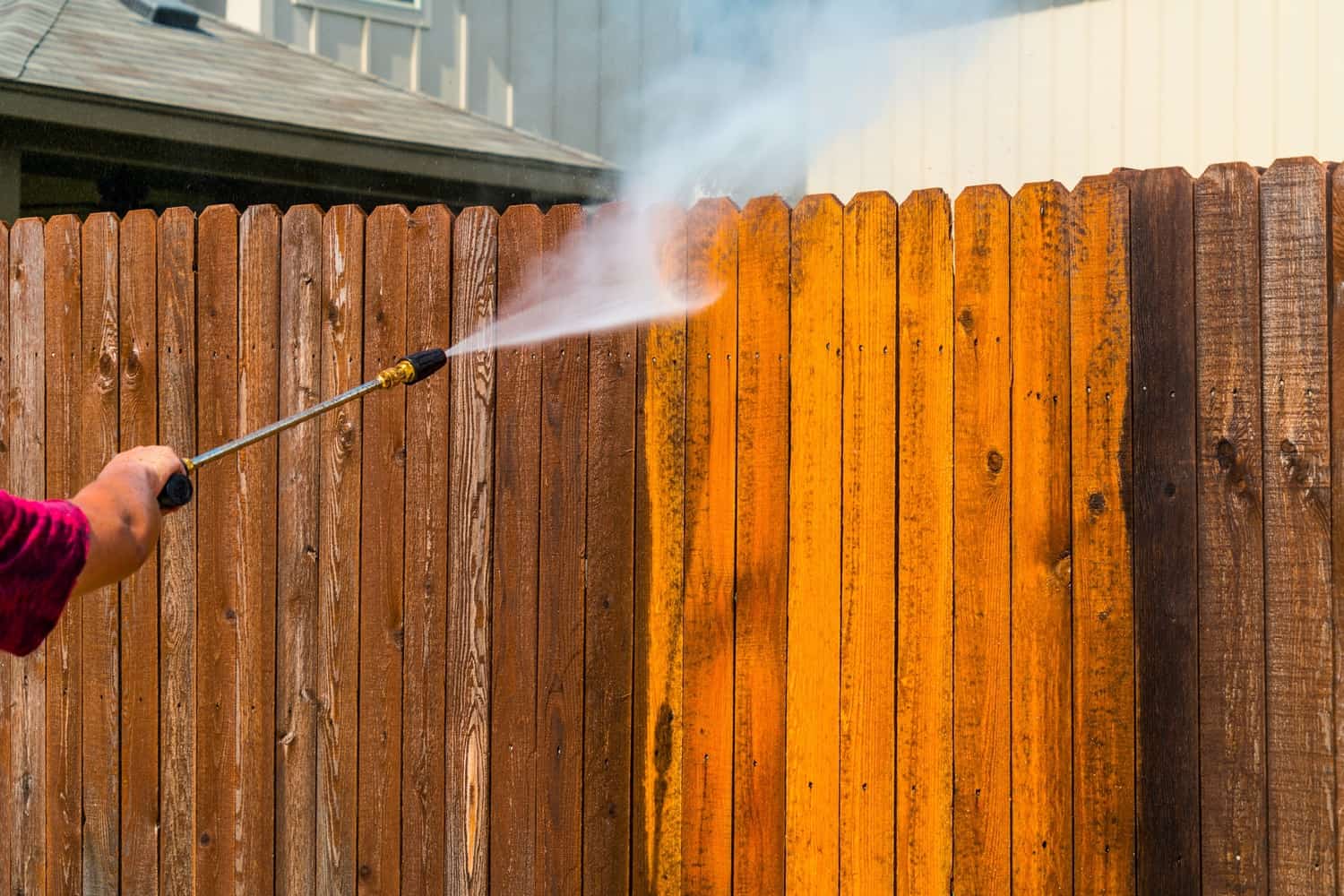 Fence Pressure Washing Service Worker pressure washing orange and brown stained vertical wood fence boards with high-pressure spray nozzle