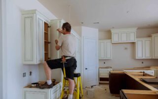 Worker installing cream-painted cabinetry with raised-panel doors in residential kitchen during renovation