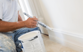 Painter's hand with brush applying white paint to interior wall trim during residential painting project.