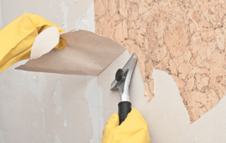 Hands in yellow gloves scraping tan textured cork wallpaper from white drywall with metal putty knife during interior