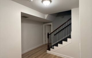 White-painted residential basement with light wood flooring, black metal stair railings, and recessed ceiling lighting.