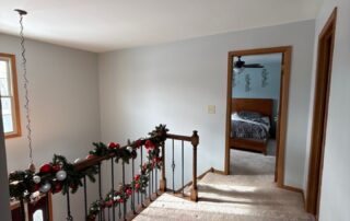 White-painted residential upstairs hallway with beige carpeting, wood-trimmed doorways, and stair railing decorated with