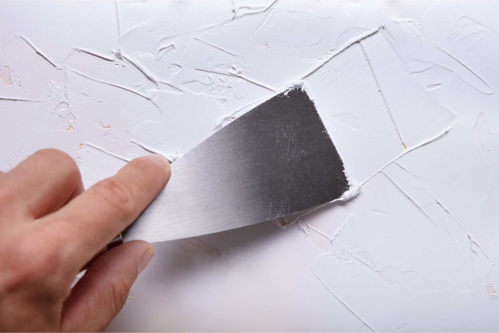 Drywall Scraping During Interior Painting Hand holding putty knife scraping textured white drywall surface during interior residential painting preparation.