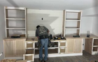 Worker installing light wood-tone cabinetry with white shiplap backsplash and open shelving in residential garage