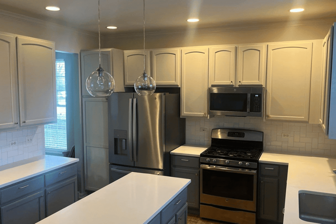 Cream-painted upper kitchen cabinets with blue lower cabinets, white countertops, stainless steel appliances, and pendant