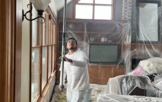Painter in white coveralls preparing interior room with plastic sheeting protection and natural wood trim for residential what to do before painters come