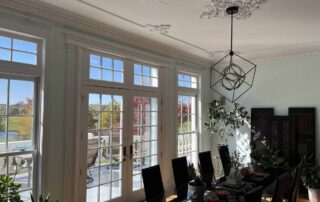 Cream-painted dining room with damaged ceiling plaster, white trim, black dining table, and large windows overlooking water.