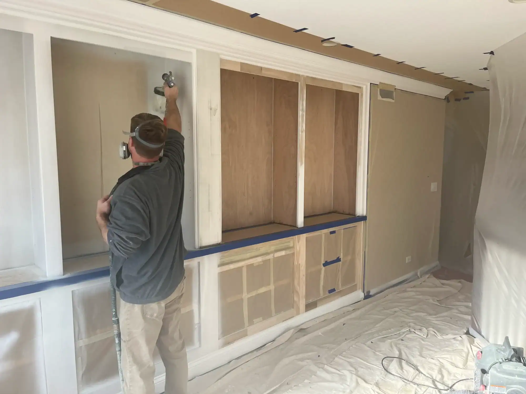 Interior Residential Painting Preparation Painter in dark shirt applying tape to white-trimmed built-in cabinetry with wood doors in beige interior room during