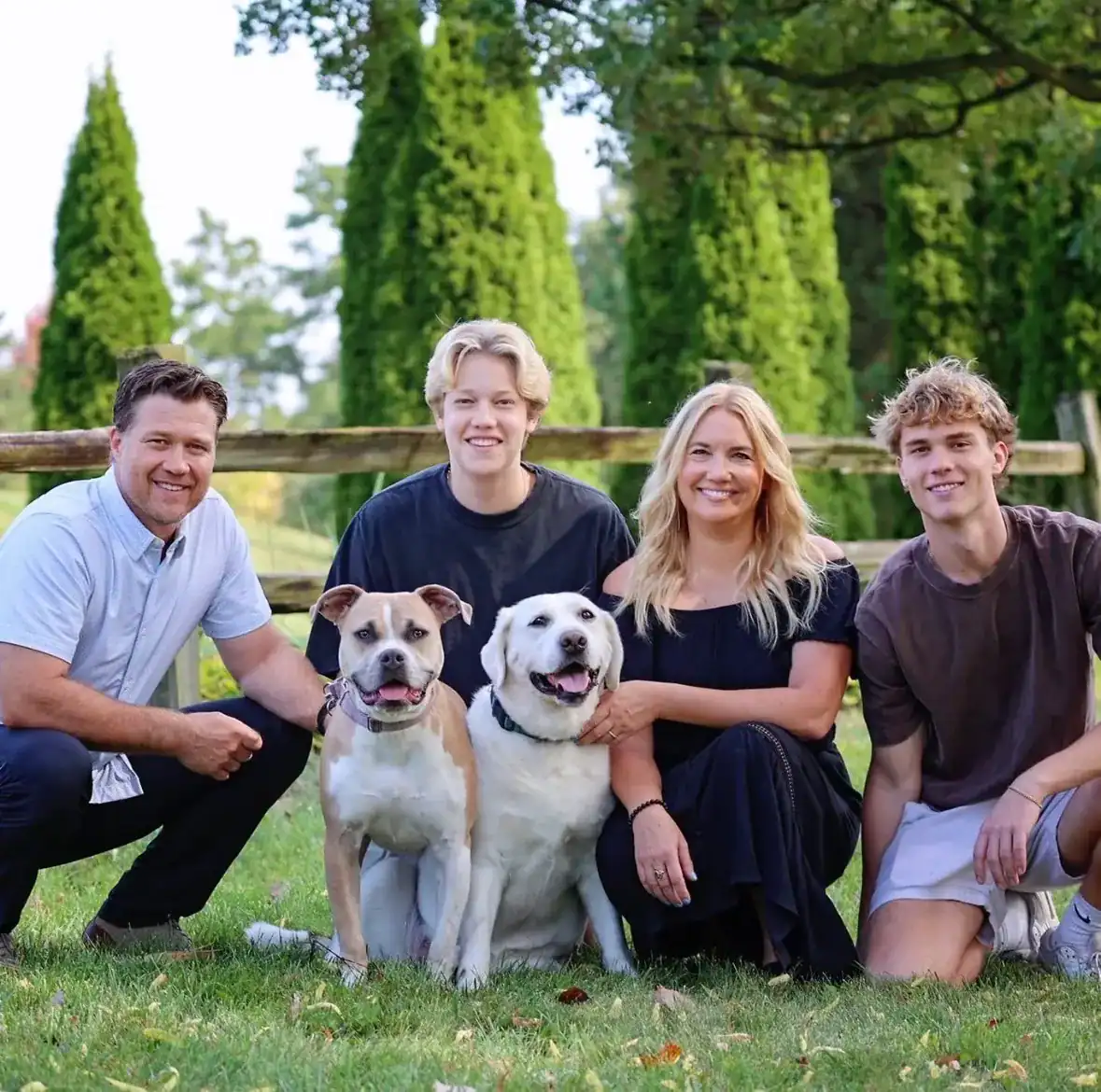 Family Photo With Dogs Family of four posing outdoors with two dogs on grass, wooden fence and evergreen trees in background.