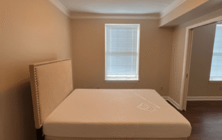 Tan-painted residential bedroom with white trim, cream upholstered bed, windows, and dark wood flooring