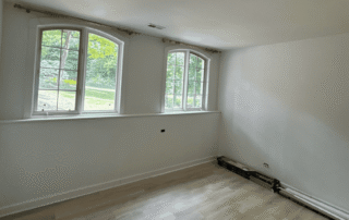 Cream-painted residential bedroom with white trim, two large windows with natural light, and light wood flooring.