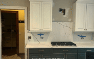 Two-tone kitchen with cream upper cabinets, dark teal lower cabinets, marble backsplash, and white countertops during