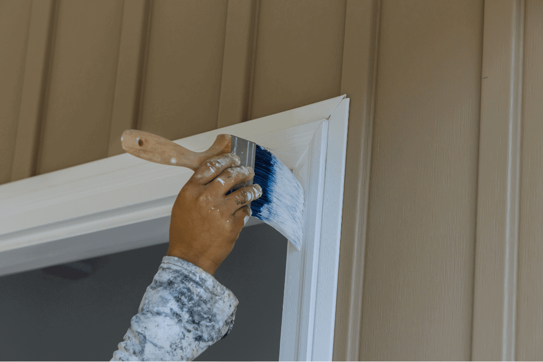 Interior Door Frame Trim Painting Painter's hand with blue brush applying white paint to interior door trim frame against beige wall.