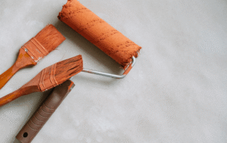Orange paint roller and two orange brushes with wooden handles arranged on white surface.