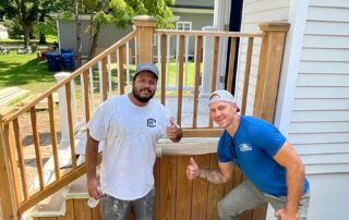 Two workers on natural wood deck with white-painted posts and railings beside cream vinyl-sided residential home