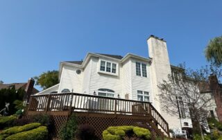 White-painted two-story residential home with stone chimney, dark wood deck railings, and horizontal vinyl siding.