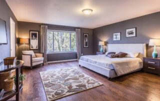 Gray-painted residential master bedroom with white upholstered bed, warm wood flooring, area rug, and large windows.
