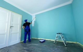 Painter in blue uniform applying turquoise paint to residential interior wall with white trim and protected flooring.