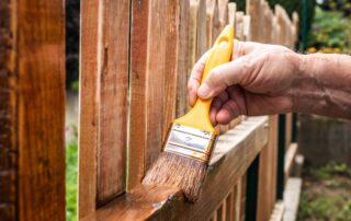 Worker applying medium brown wood stain to exterior residential fence boards with yellow-handled paintbrush.