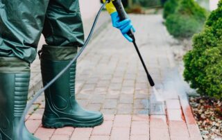 Worker in green boots and blue gloves pressure washing red and tan brick pavers with visible dirt removal.