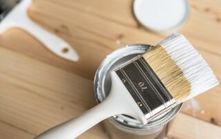 Paint brush with white handle and natural bristles resting across open can of white paint on wooden surface.