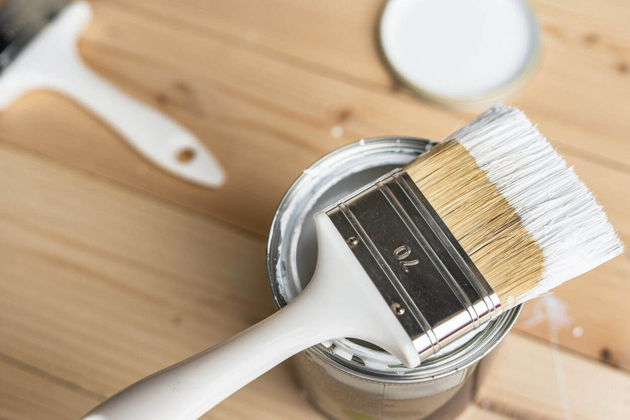 Paint Brush in White Paint Can Paint brush with white handle and natural bristles resting across open can of white paint on wooden surface.