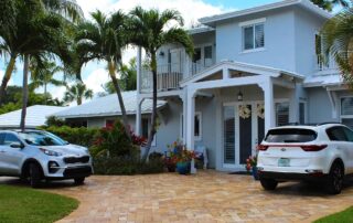 Light blue two-story residential home exterior with white trim, covered porch, and brick paver driveway.