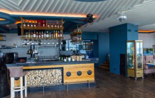 Deep teal-blue painted bar interior with white corrugated metal ceiling, wood counter, and ambient orange accent lighting