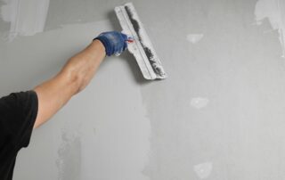 Worker in blue glove sanding white drywall surface with sanding block during interior painting preparation.