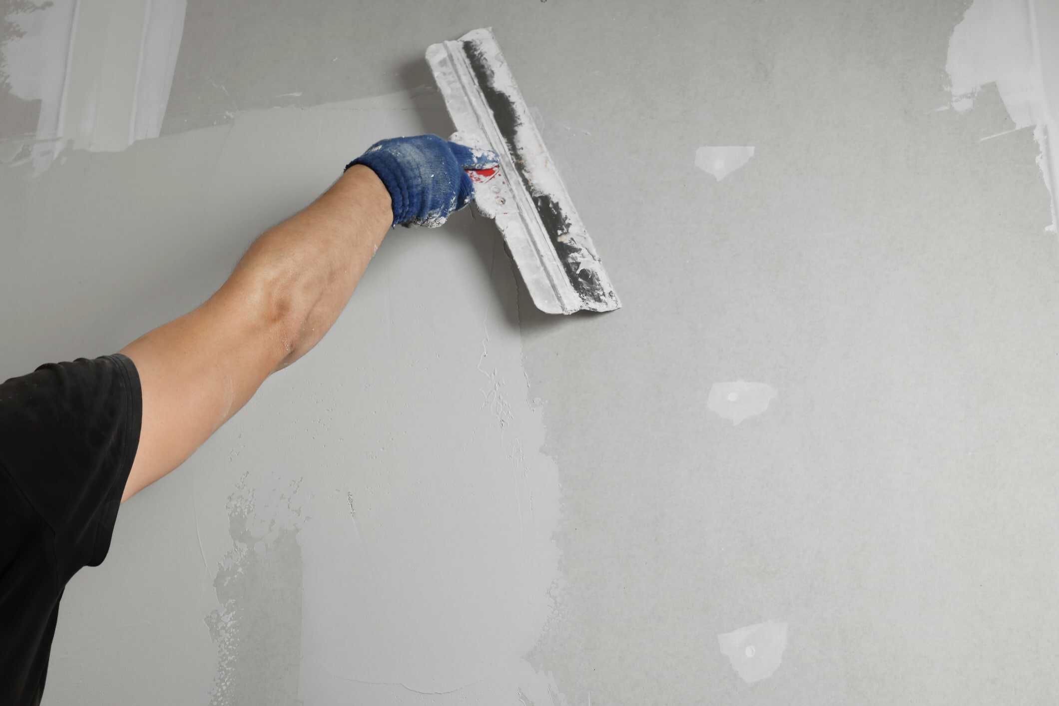 Drywall Sanding Preparation Worker in blue glove sanding white drywall surface with sanding block during interior painting preparation.