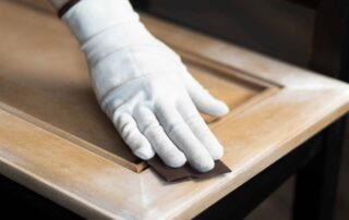 Hand in white cotton glove applying dark brown stain to natural wood door trim with brush during interior residential