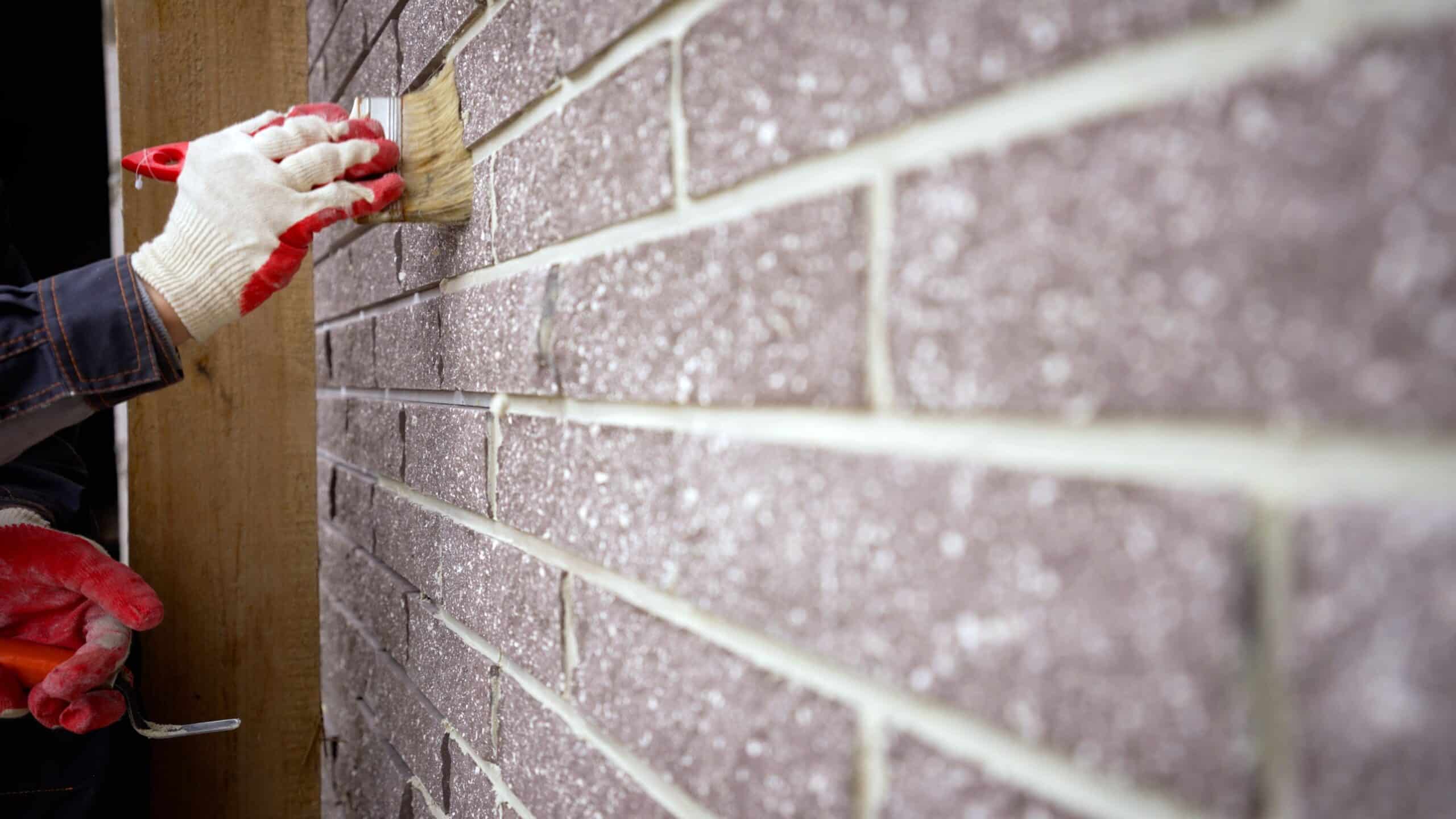 Exterior Brick Wall Painting Painter in white gloves applying red paint to gray brick exterior wall with paintbrush during residential painting.