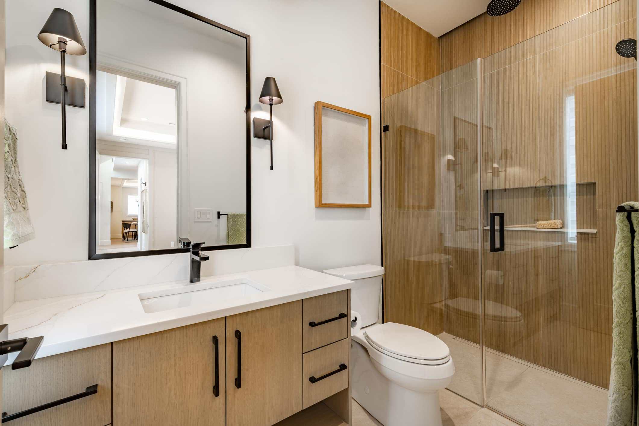 Modern Bathroom With White Vanity White quartz countertop with natural wood cabinetry, black-framed mirror, and glass shower enclosure in residential bathroom.