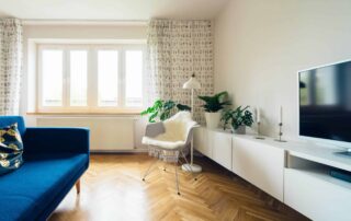 Cream-painted residential living room with white trim, herringbone wood flooring, blue sofa, and large windows with sheer
