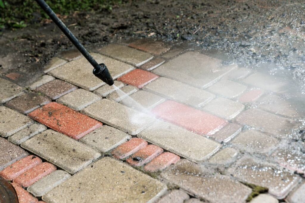 Pressure Washing Pavers Pressure washer spray cleaning alternating red and tan brick pavers with visible dirt and moss removal.