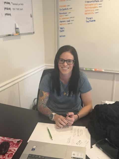 Woman with dark hair and glasses wearing gray shirt seated at desk with laptop and notepad in office.
