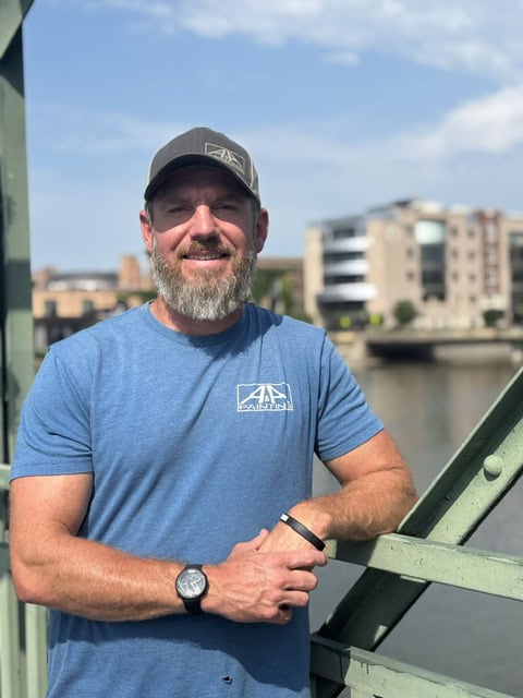 Man in blue t-shirt and gray cap smiling at camera on waterfront bridge with city buildings in background.