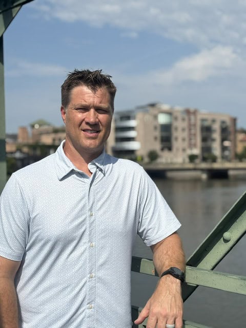 Man in light blue short-sleeve button-up shirt smiling at camera on outdoor bridge with waterfront cityscape background.