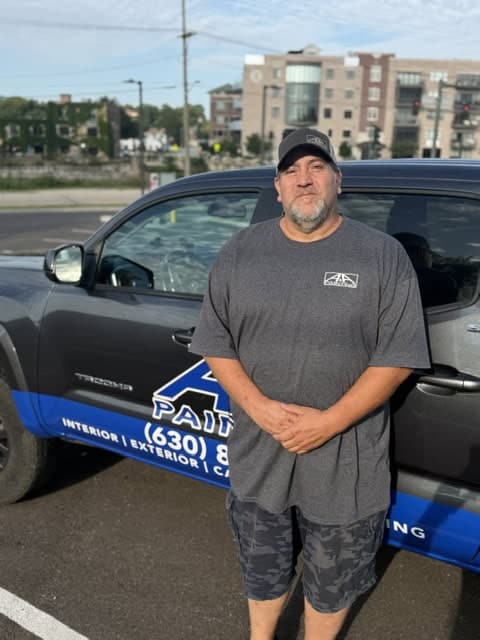 Man in gray shirt and cap standing beside blue and black company work vehicle in parking lot.