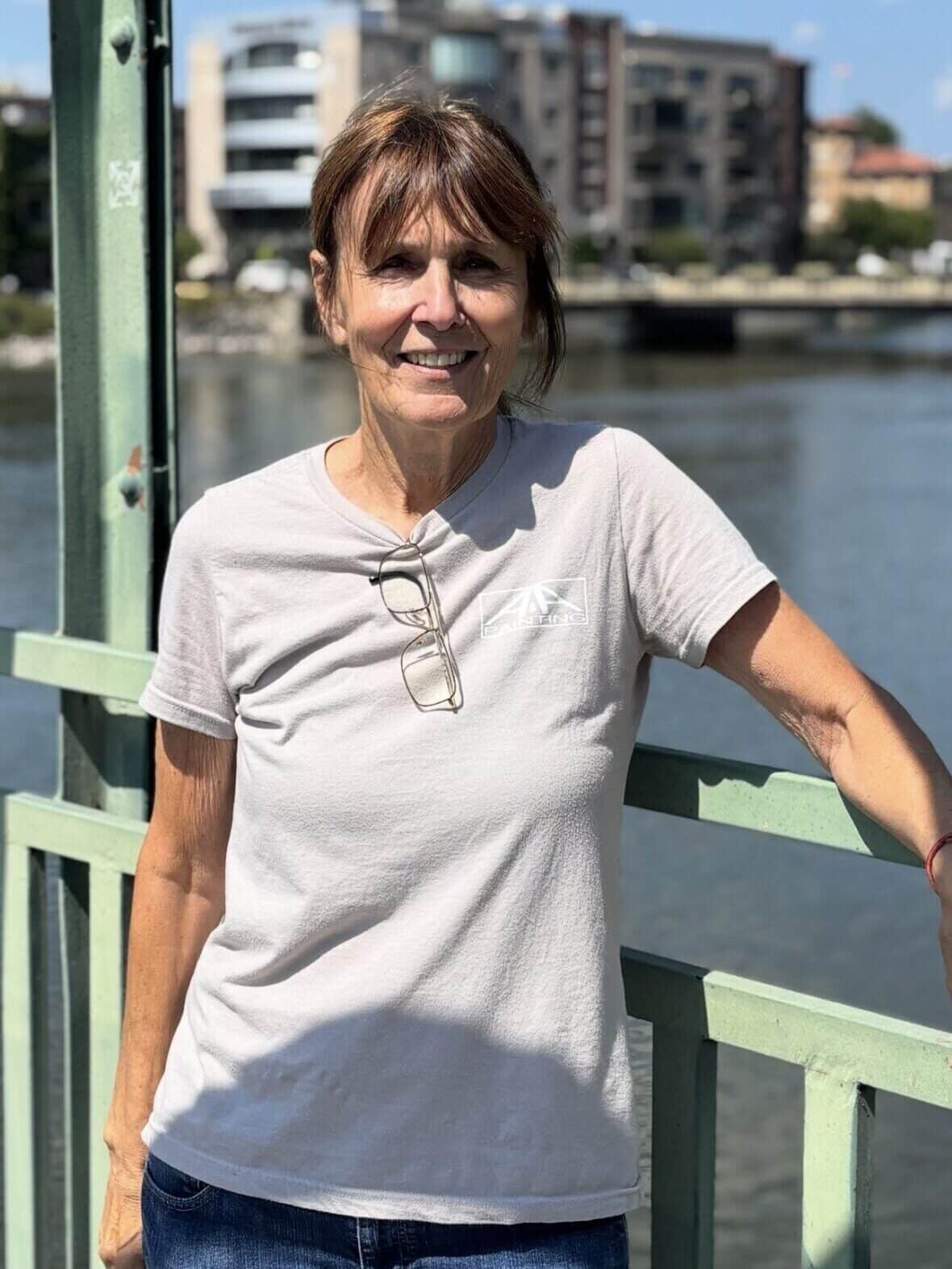 Woman in white t-shirt smiling at camera on green metal bridge railing over water.