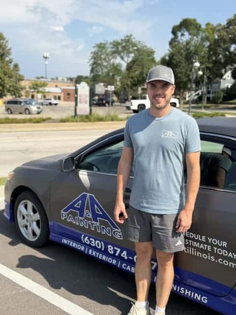 Man in light gray t-shirt and white cap standing beside company work vehicle in parking lot.